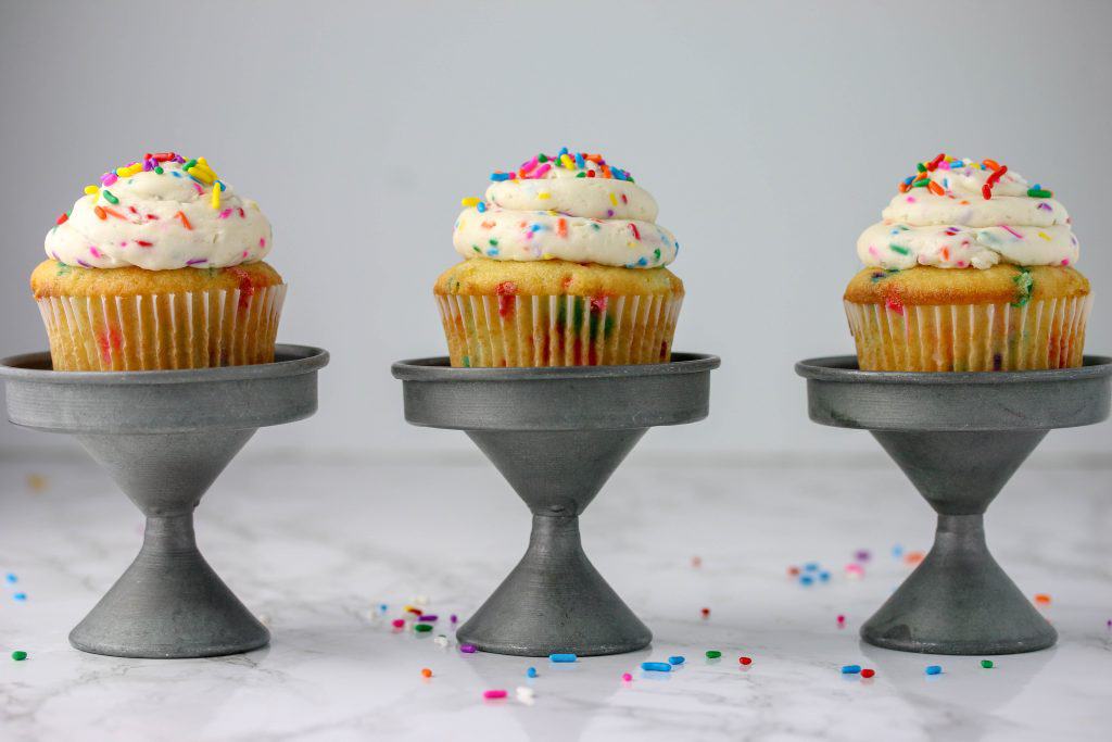 three funfetti cupcakes with cake batter frosting on individual cupcake stands 