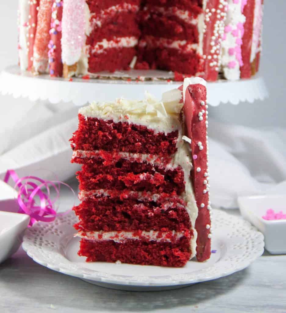 slice of 4 layer red velvet cake with cake on a cake stand behind it