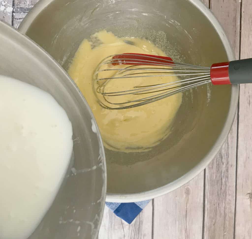 Milk being poured into egg yolks in a metal bowl with a whisk