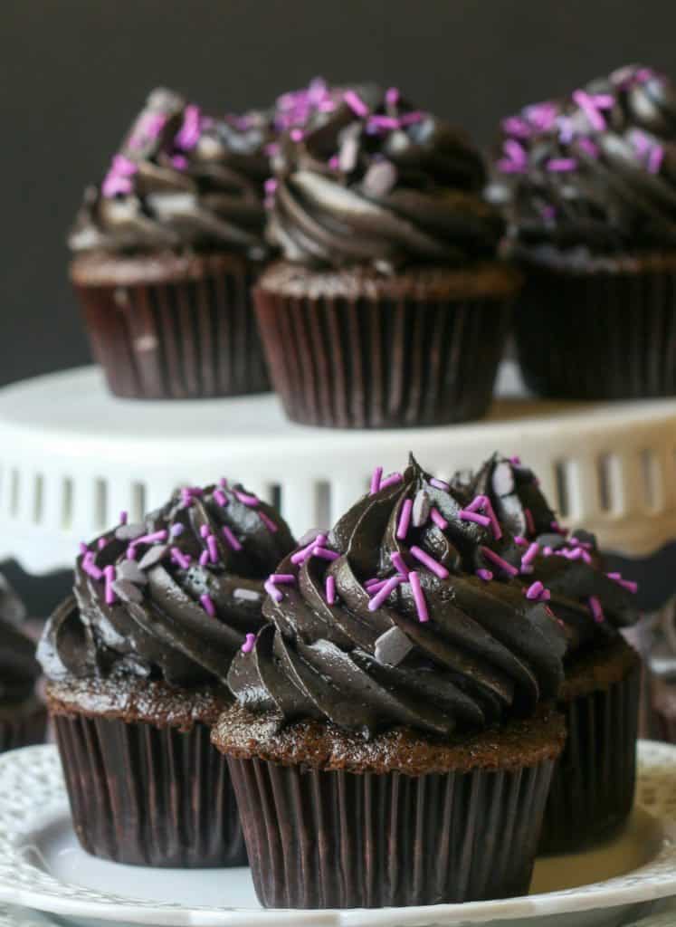 three black icing cupcakes with purple sprinkles on a plate with cupcakes on a cake stand behind it