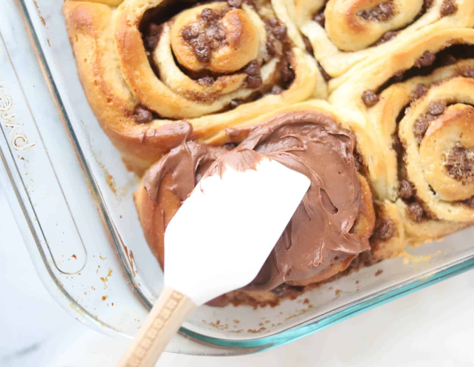 chocolate cream cheese frosting being spread onto baked cinnamon rolls