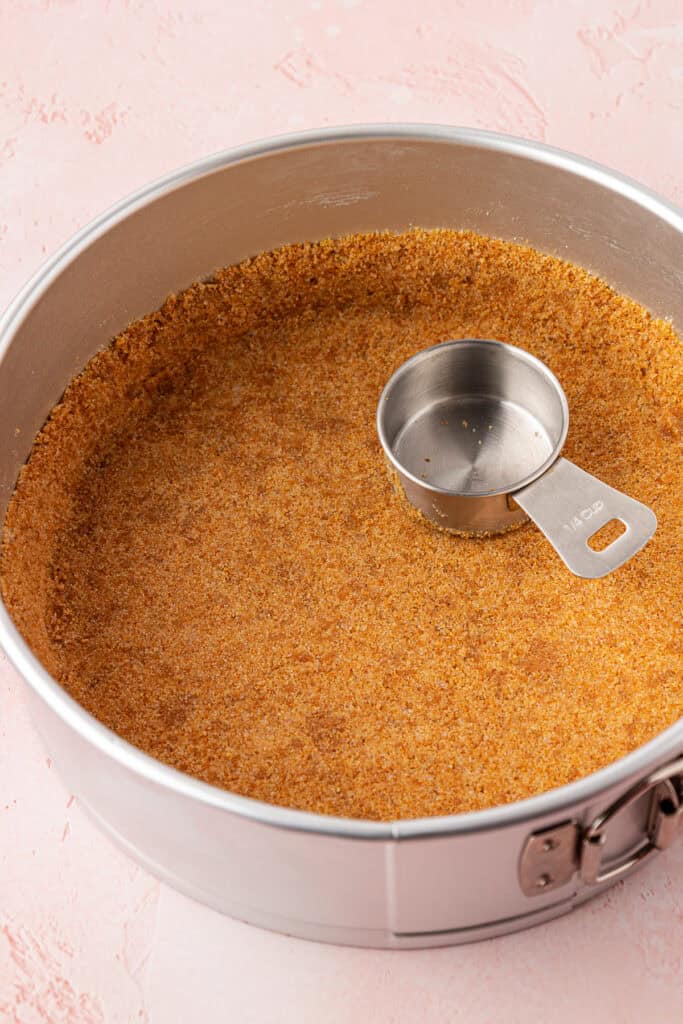 graham cracker crust in a in a spring form pan pressed down by a measuring cup