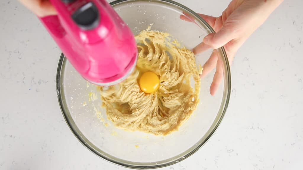 pink hand mixer mixing an egg into creamed butter/sugar mixture