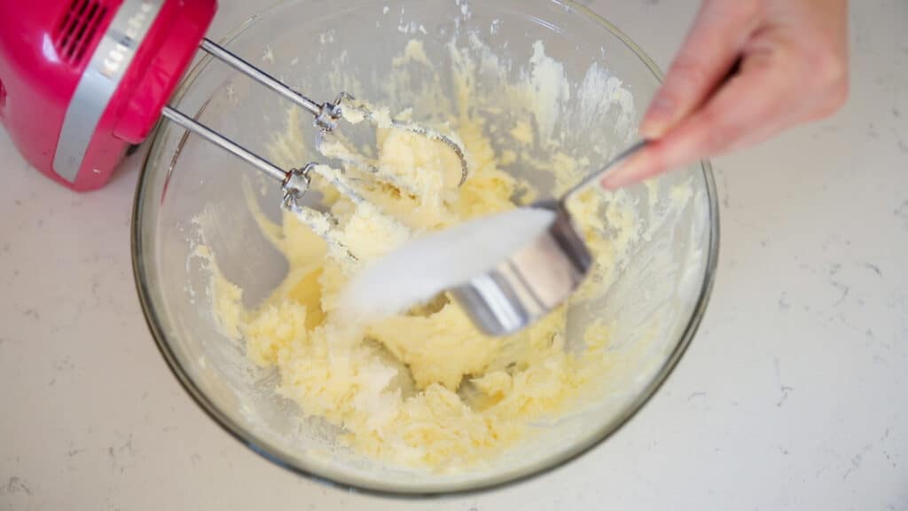 metal measuring cup of sugar added to a bowl of butter with a pink hand mixer in it