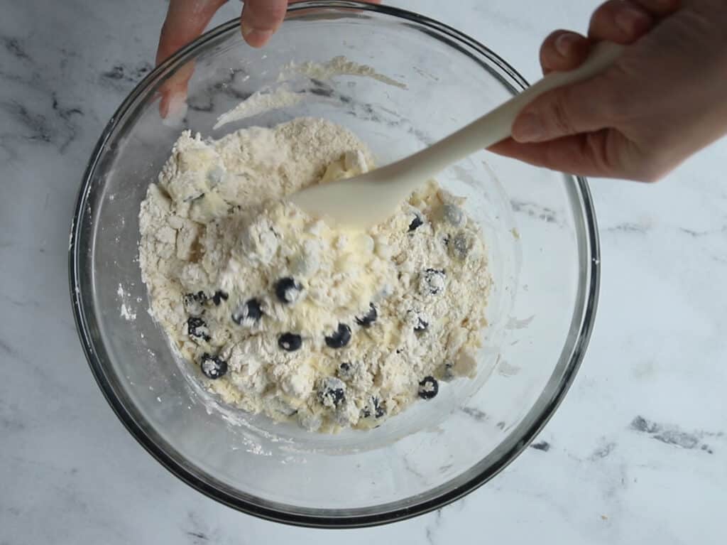 hand stirring with a spatula blueberries and lemon zest into bowl of dry ingredients