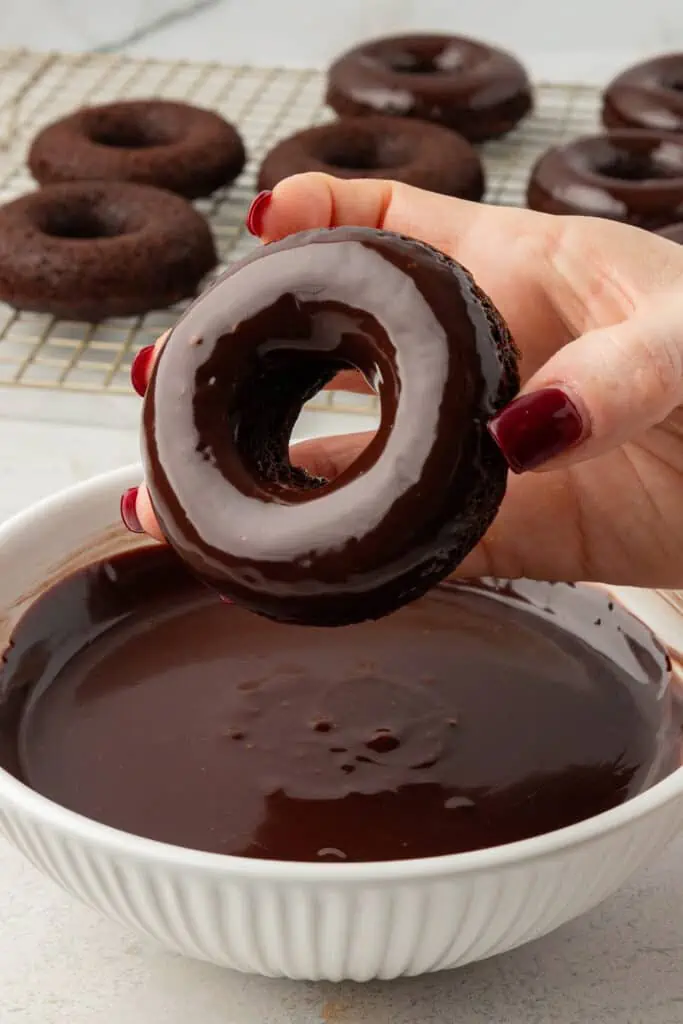 hand dipping baked chocolate donut in a bowl of chocolate glaze