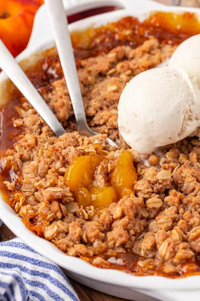 peach crisp with two spoons in it and a scoop of vanilla ice cream
