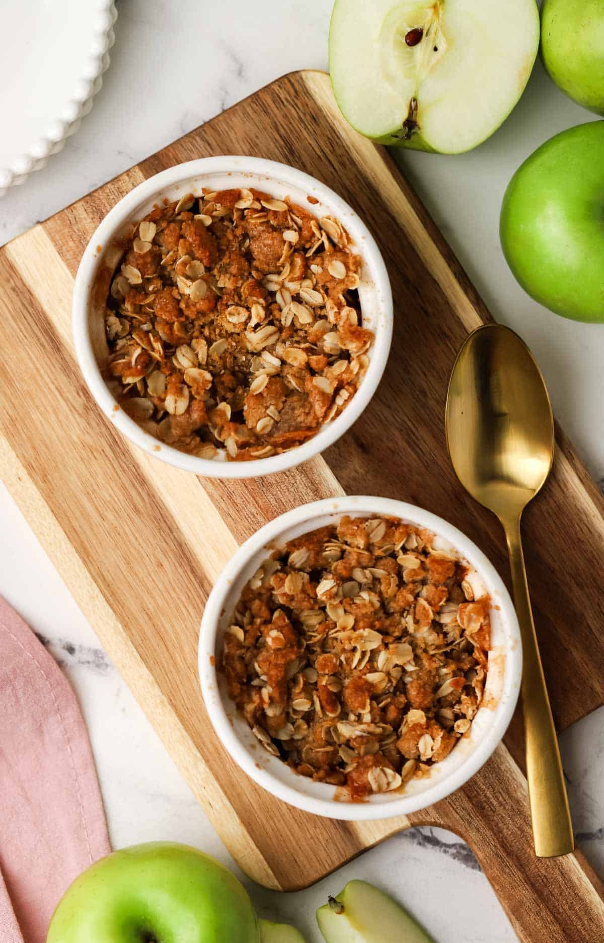 two baked apple crisp ramekins on a small cutting board